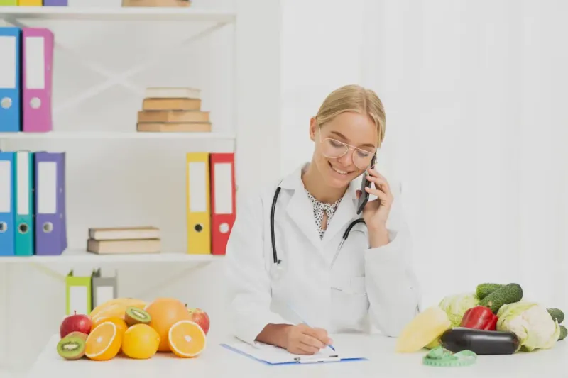 Doctora sonriendo mientras habla por teléfono rodeada de frutas y verduras saludables.