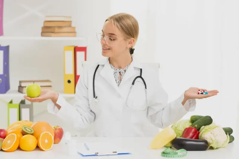 Un médico sostiene una manzana en una mano y pastillas en la otra, rodeado de frutas, verduras y herramientas médicas sobre un escritorio.